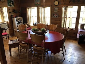 A wooden dining room with a round table and chairs, a red tablecloth, and a bowl of greenery on the table.
