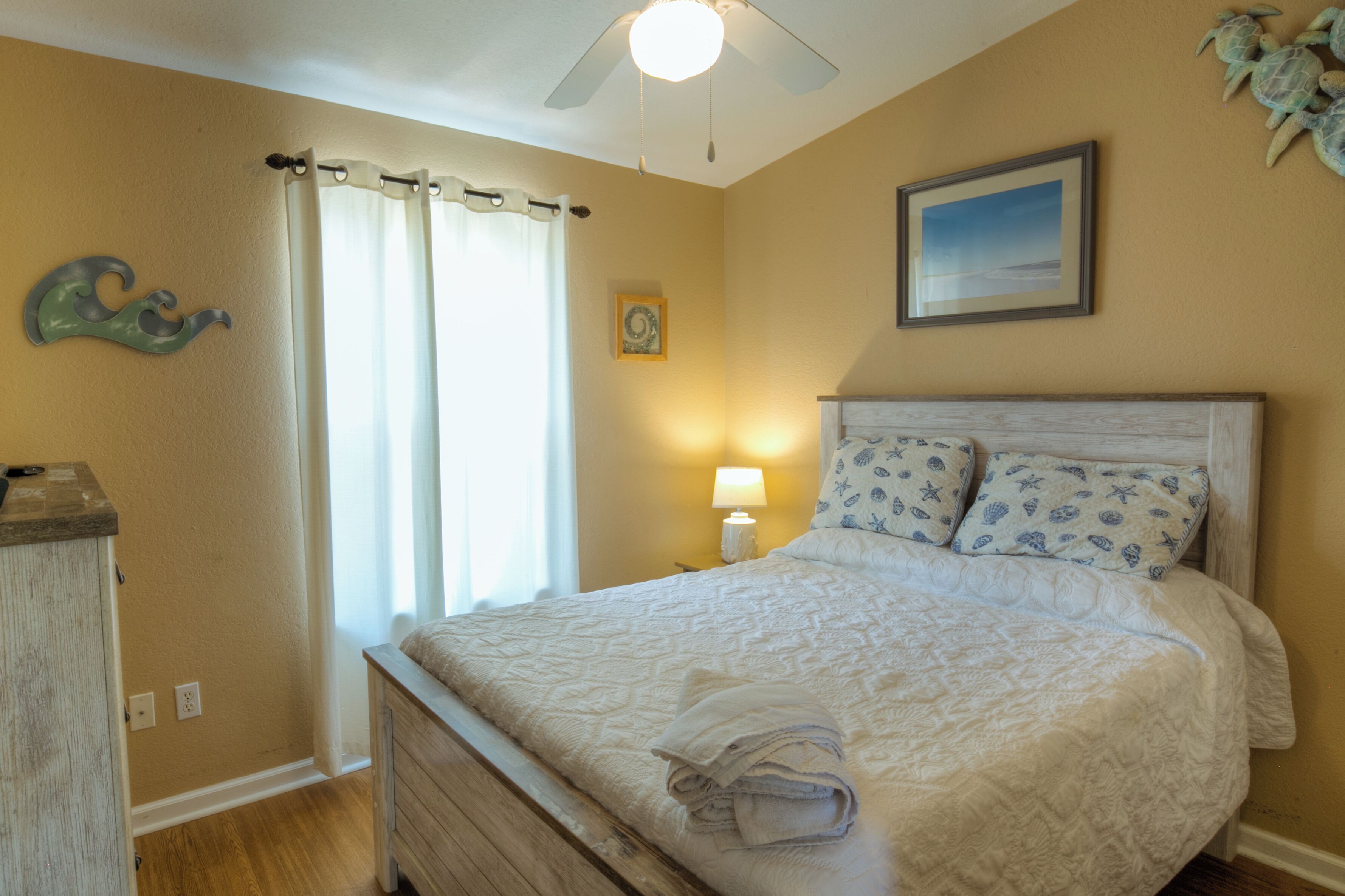 A bedroom with a bed, curtains, a ceiling fan, a nightstand with a lamp, and a wall-mounted picture.
