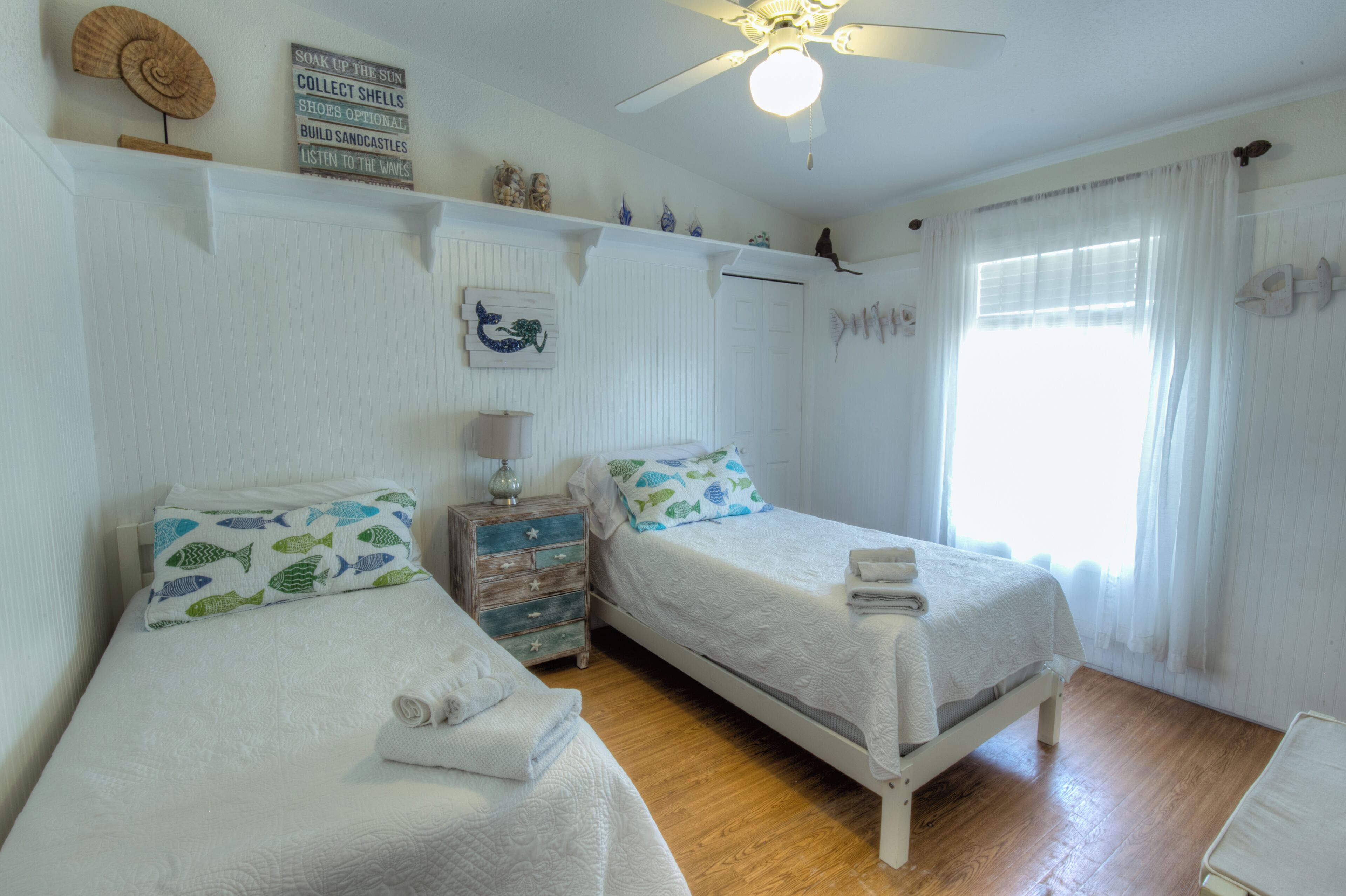 A bedroom with two beds, a ceiling fan, a window with curtains, and a wall with decorative items.