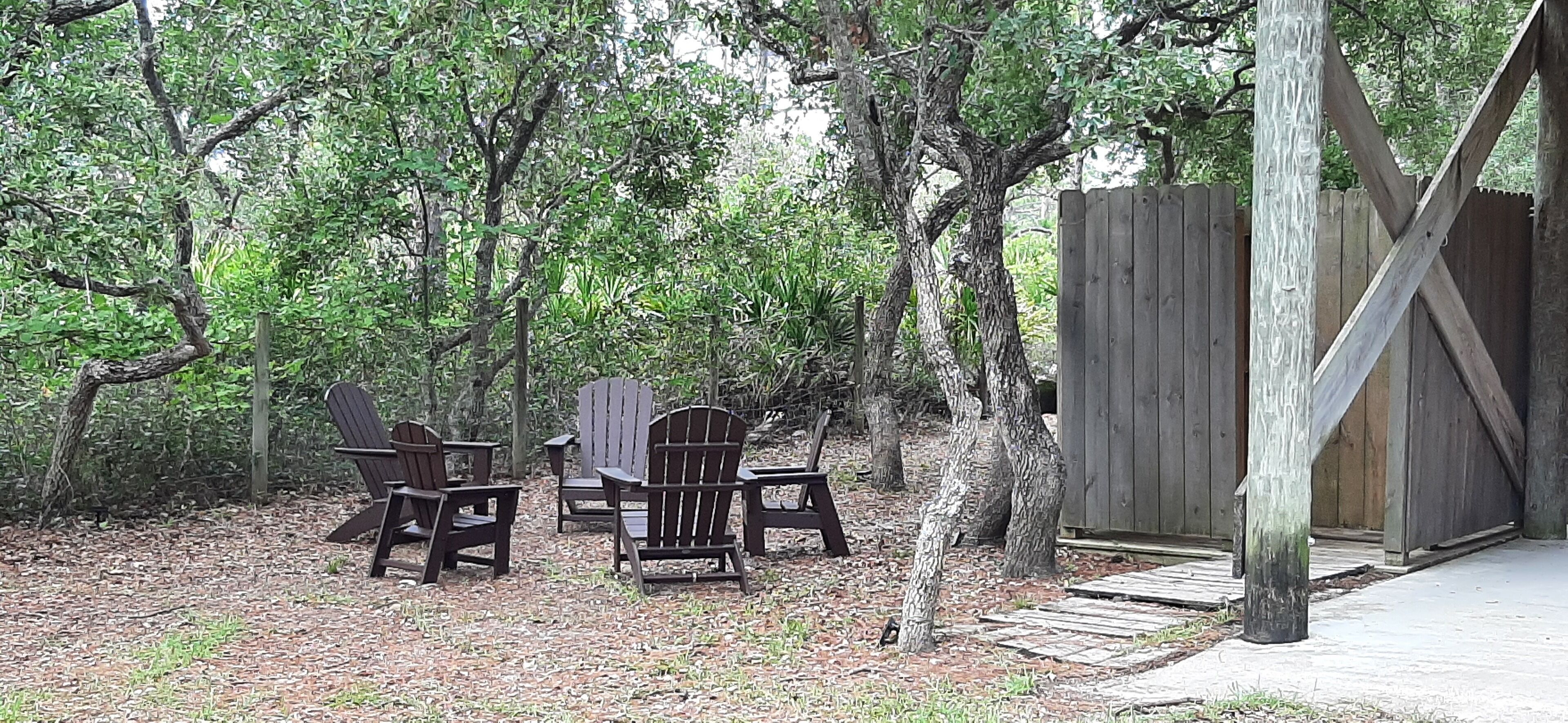 An outdoor seating area with wooden chairs and tables, surrounded by trees and a wooden fence.