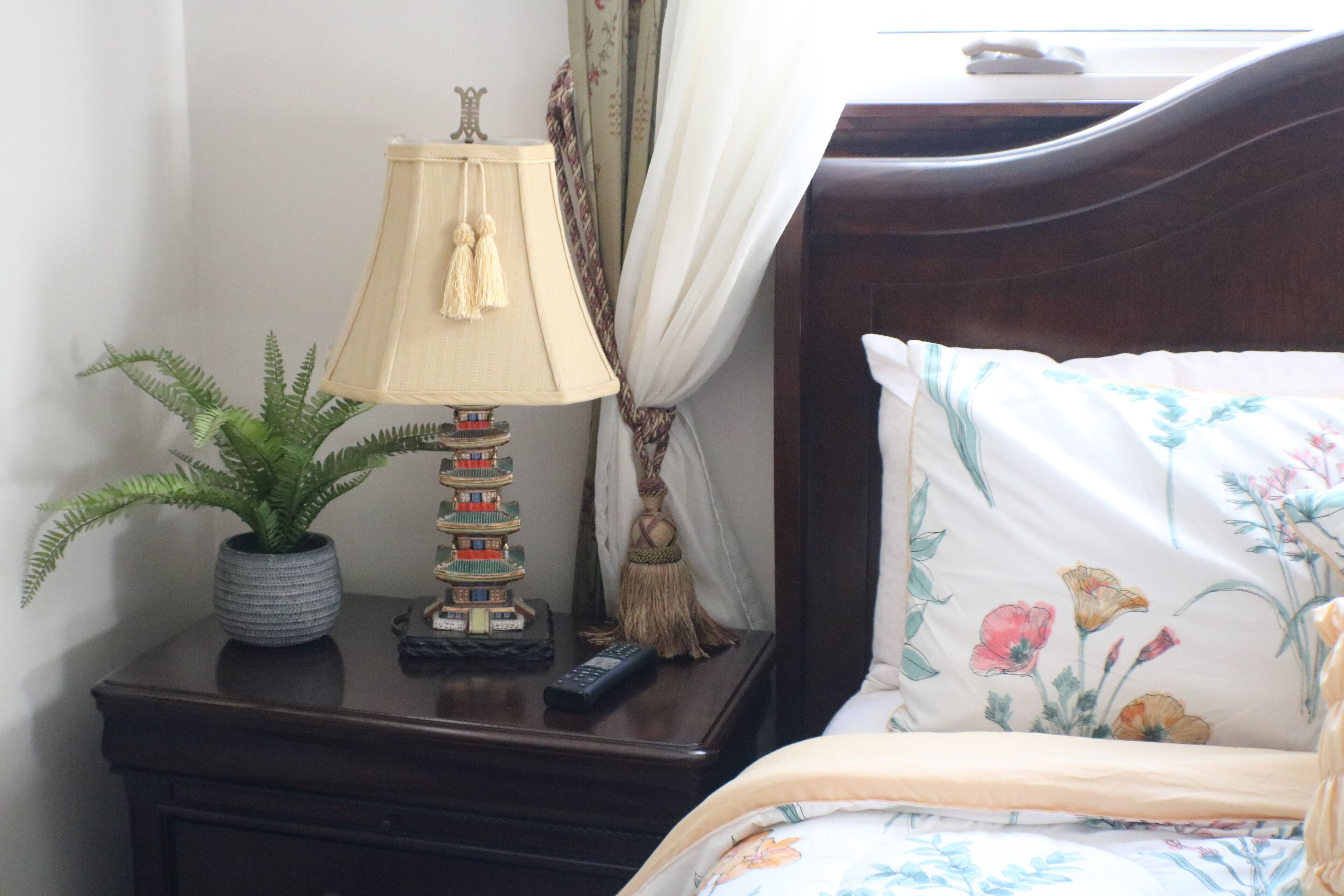 Una cama bien tendida con ropa de cama floral, una lámpara de noche, una pequeña planta en maceta y un control remoto sobre una mesita de noche de madera.