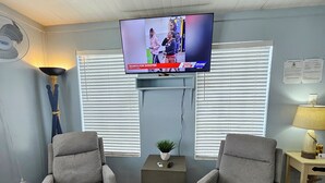 A living room with two grey armchairs, a television mounted on the wall, a small table with a plant, and a window with blinds. - Fisherman's Get-A-Way! Cozy waterfront retreat with access to Lake Okeechobee! (Okeechobee)