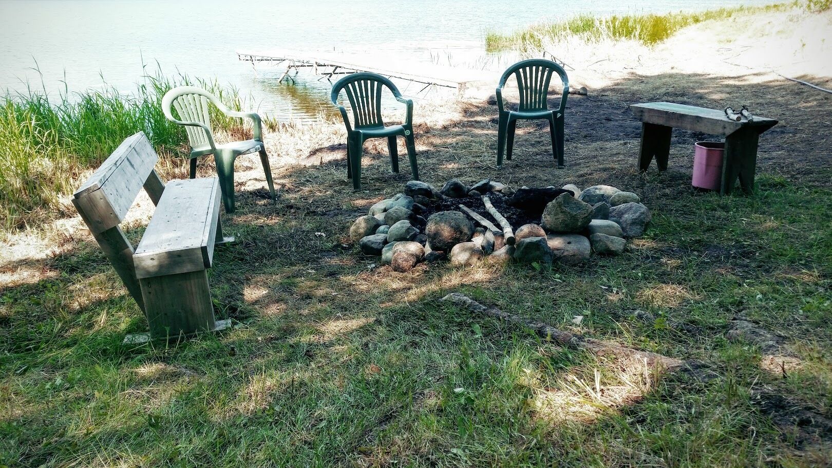 A lakeside picnic area with a fire pit, benches, and chairs.