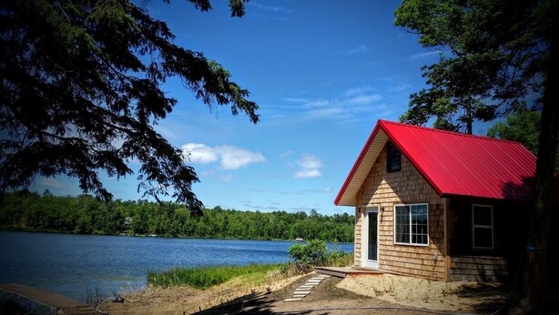 A small cabin with a red roof by a lake, surrounded by trees.