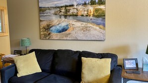 Una sala de estar con un cuadro grande de un paisaje de géiser en la pared, un sofá oscuro con cojines amarillos, una lámpara y una pequeña mesa con un marco de fotos.