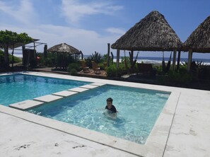 A swimming pool with a child in the water, surrounded by lounge chairs and thatched umbrellas, with a view of the ocean. - Capricho Beach House (Jujutla)
