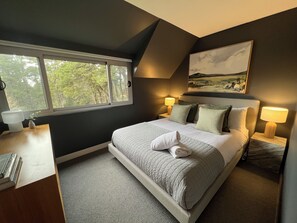 A bedroom with a bed, two bedside lamps, a window with a view of trees, a painting on the wall, and a small plant on the bedside table. - Maple View Medlow Bath, Blue Mountains (Medlow Bath)