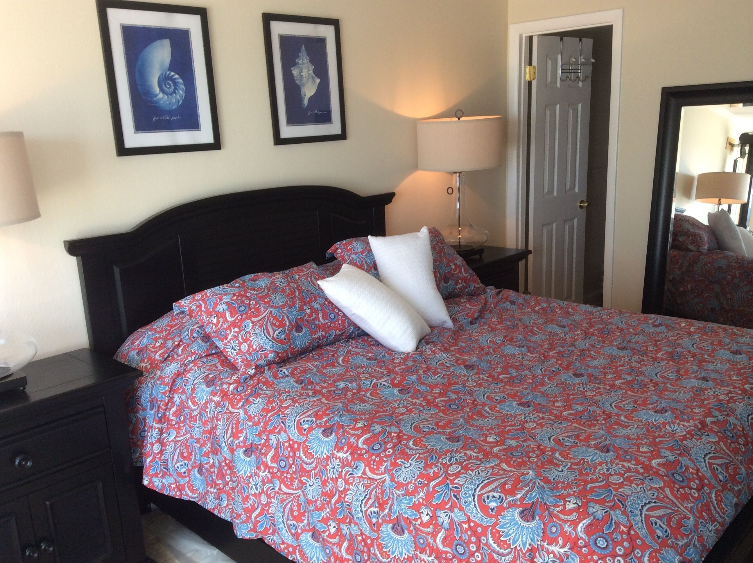 A neatly made bed with a patterned bedspread, two framed pictures on the wall, a bedside lamp, and a door with a mirror.