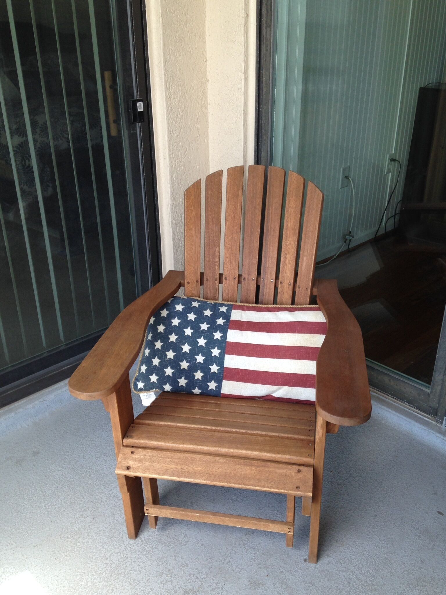 Una silla Adirondack de madera con un cojín con la bandera estadounidense en un porche de concreto.