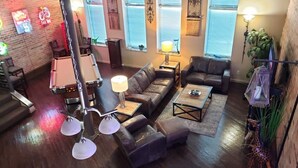 A modern living room with a sofa set, coffee table, and a pool table. There are windows with blinds, a brick wall, and a staircase leading to another level. - Up to 50% off for limited time! Professionally Cleaned, Pool & Foosball Table (Nashville)