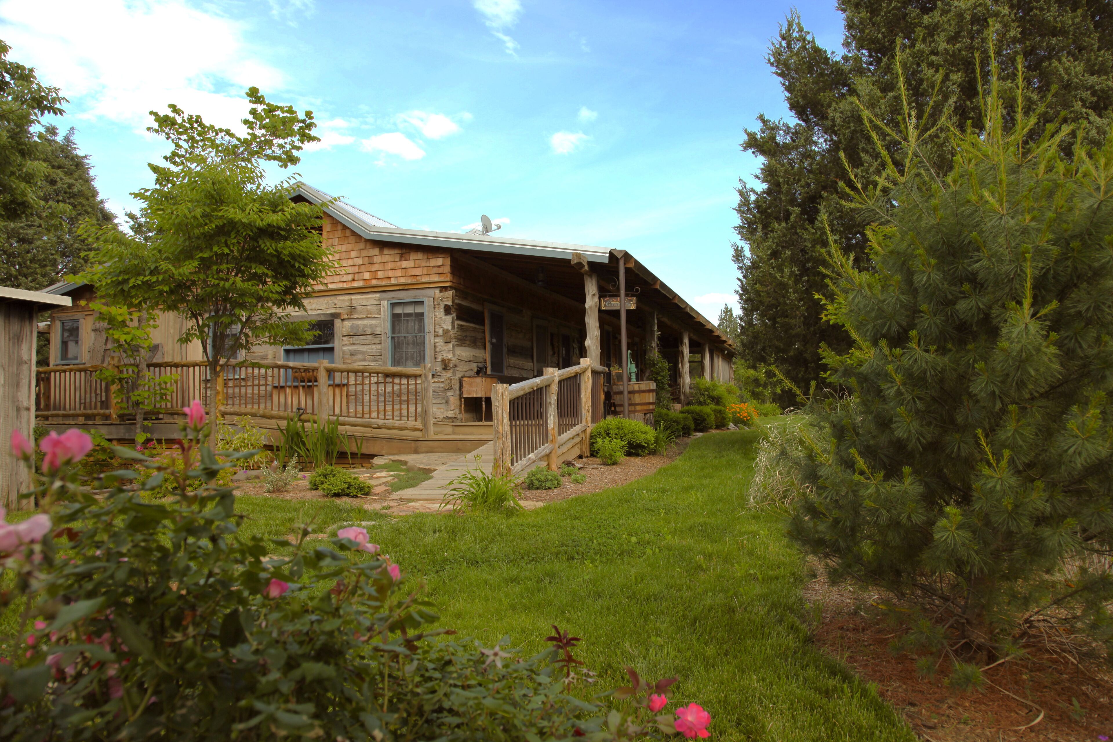 A rustic log cabin with a porch, surrounded by a well-maintained garden and tall pine trees.