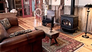 A cozy living room with a leather sofa, a small wooden table, a traditional spinning wheel, and a wood-burning stove.
