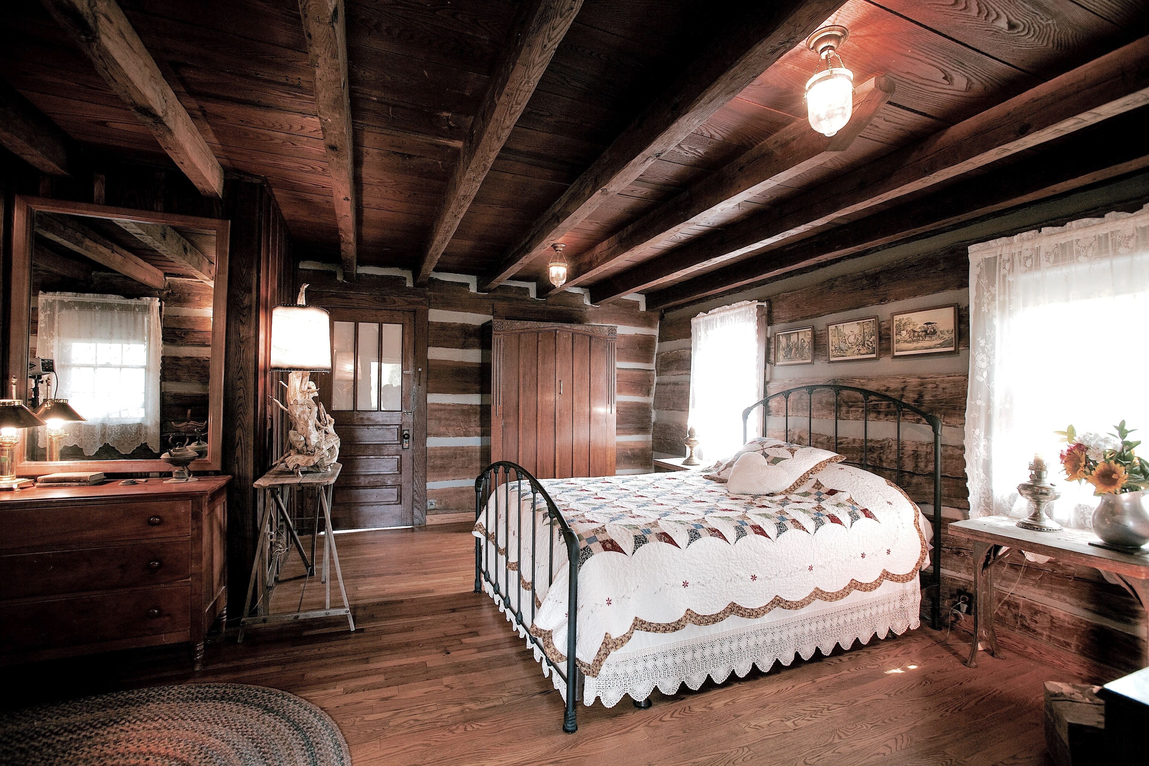 A rustic wooden cabin bedroom with a bed, dresser, mirror, and a small table with a lamp.