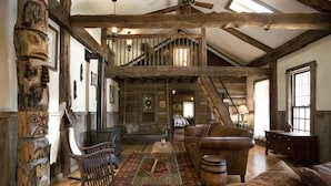 A rustic interior with exposed wooden beams, a loft area, a leather sofa, a rocking chair, and a wooden barrel.