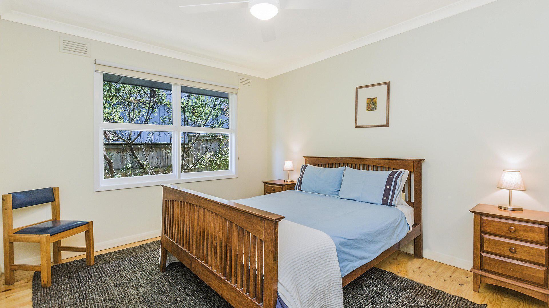 A bedroom with a wooden bed, a chair, a nightstand, and a window with a view of trees.
