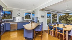 Eine moderne Küche mit Kochinsel, einem Essbereich mit Holztisch und blauen Stühlen sowie großen Fenstern mit Blick ins Grüne.