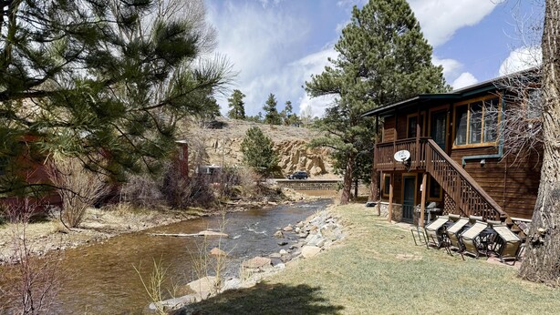 A wooden cabin with outdoor seating by a river, surrounded by trees and a rocky hillside.