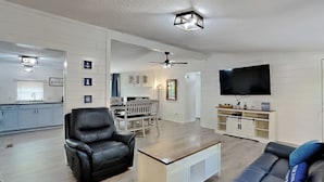 A modern living room with a black leather sofa, a white coffee table, and a black leather chair. There is a dining area with a white table and chairs in the background. A flat-screen TV is mounted on the wall, and a ceiling fan is visible. - Where Pool Days Steal the Show | 8 ppl | IC13 (Put-in-Bay)