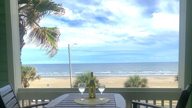 A balcony with a table and two chairs, overlooking a beach with the ocean and a cloudy sky.