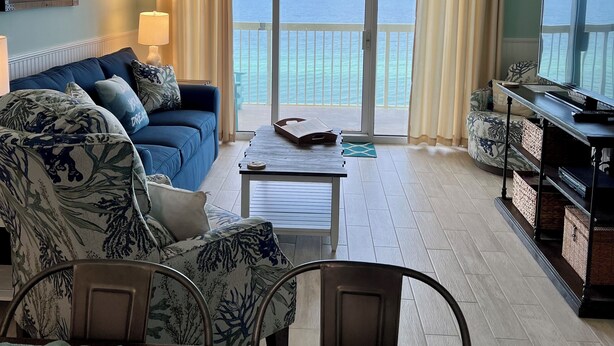 Una sala de estar con un sofá azul, una mesa de centro y un mueble para la televisión. Hay un ventanal con cortinas, un ventilador de techo y vistas al océano.