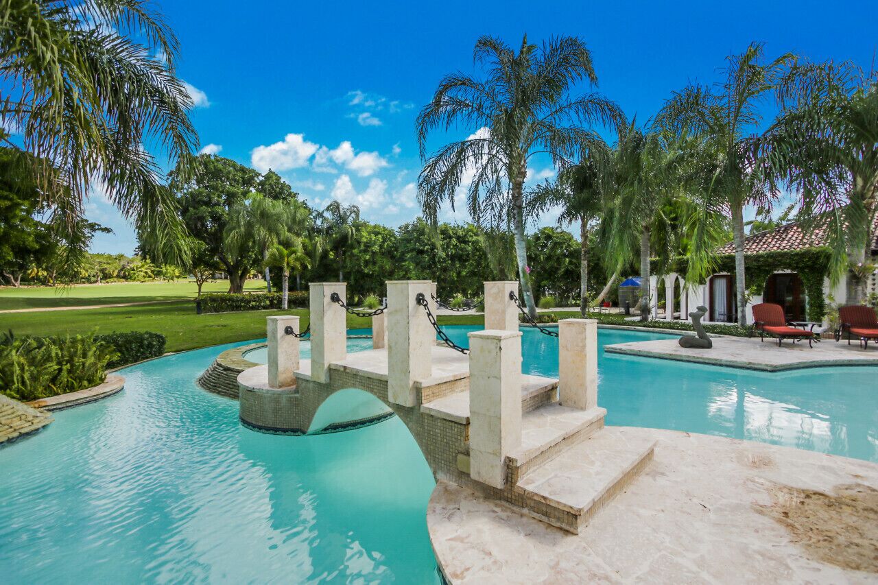 A swimming pool with a bridge, palm trees, and a building with a red roof.