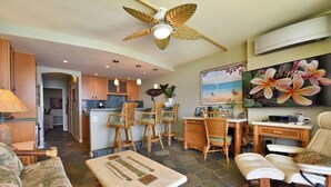 Un vaste espace de vie comprenant un canapé, une table basse, un coin repas avec des chaises, une cuisine avec des meubles en bois et un ventilateur de plafond.