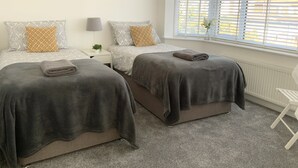 Quarto de hotel com duas camas, cada uma com um cobertor cinza e um travesseiro estampado com padrão geométrico. Há uma pequena mesa de cabeceira com uma lâmpada e uma planta. O quarto possui uma janela grande com persianas, e um radiador é visível na parede.