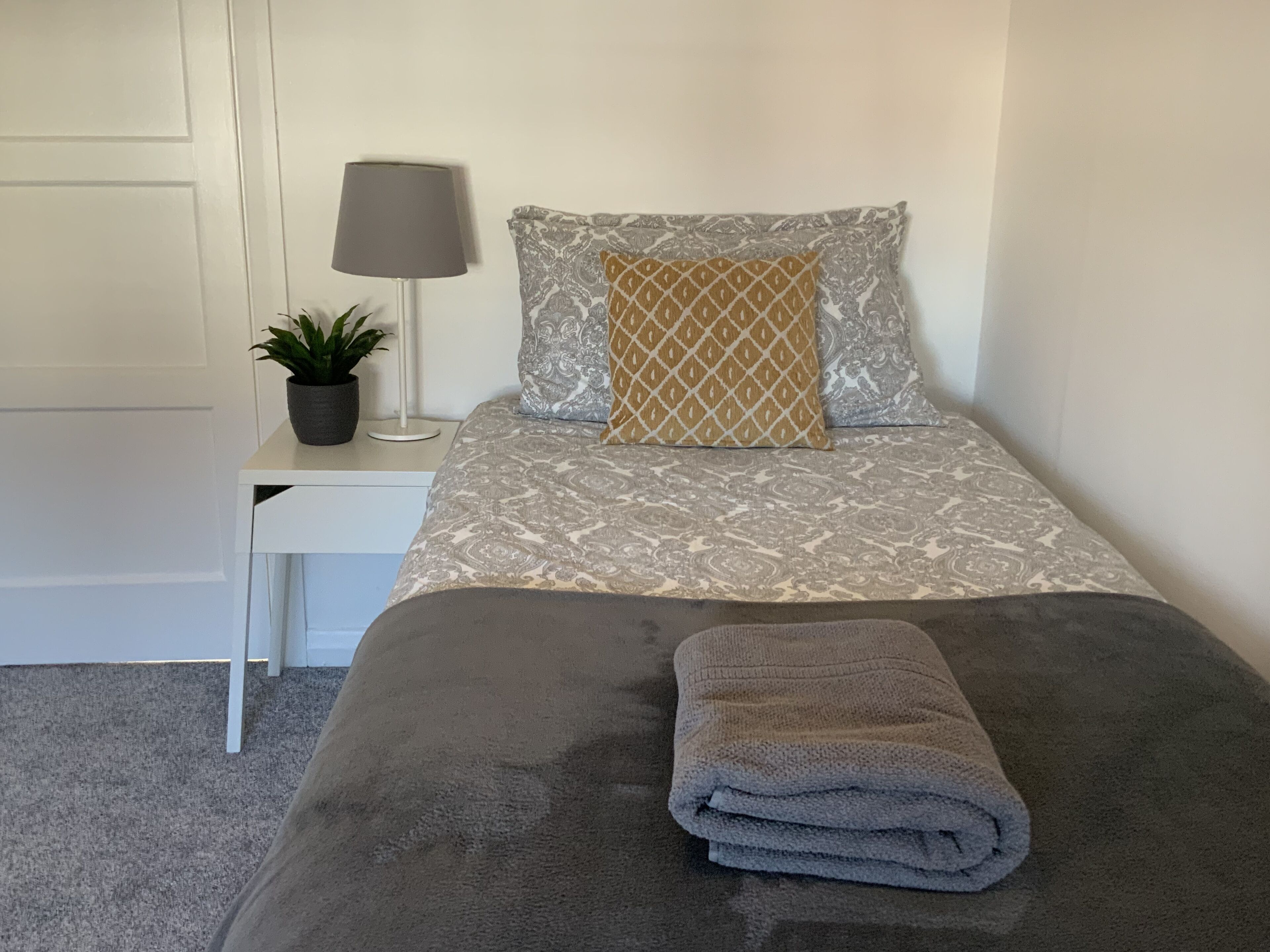 A neatly made bed with a grey blanket and a folded grey towel, a bedside table with a lamp and a potted plant, and a patterned pillow.