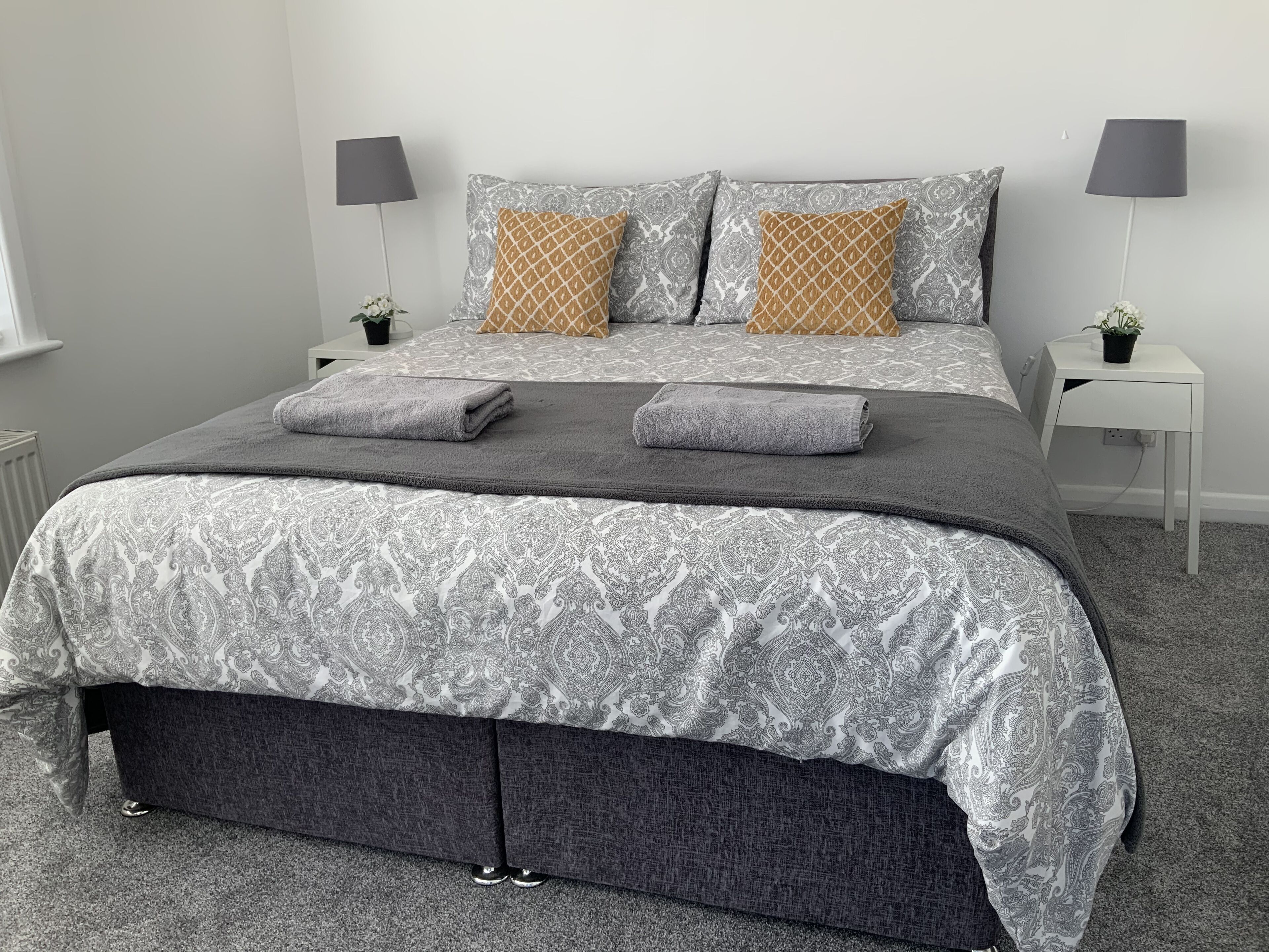 A neatly made bed with grey and white patterned bedding, two bedside tables with lamps, and a small potted plant on one of the tables.
