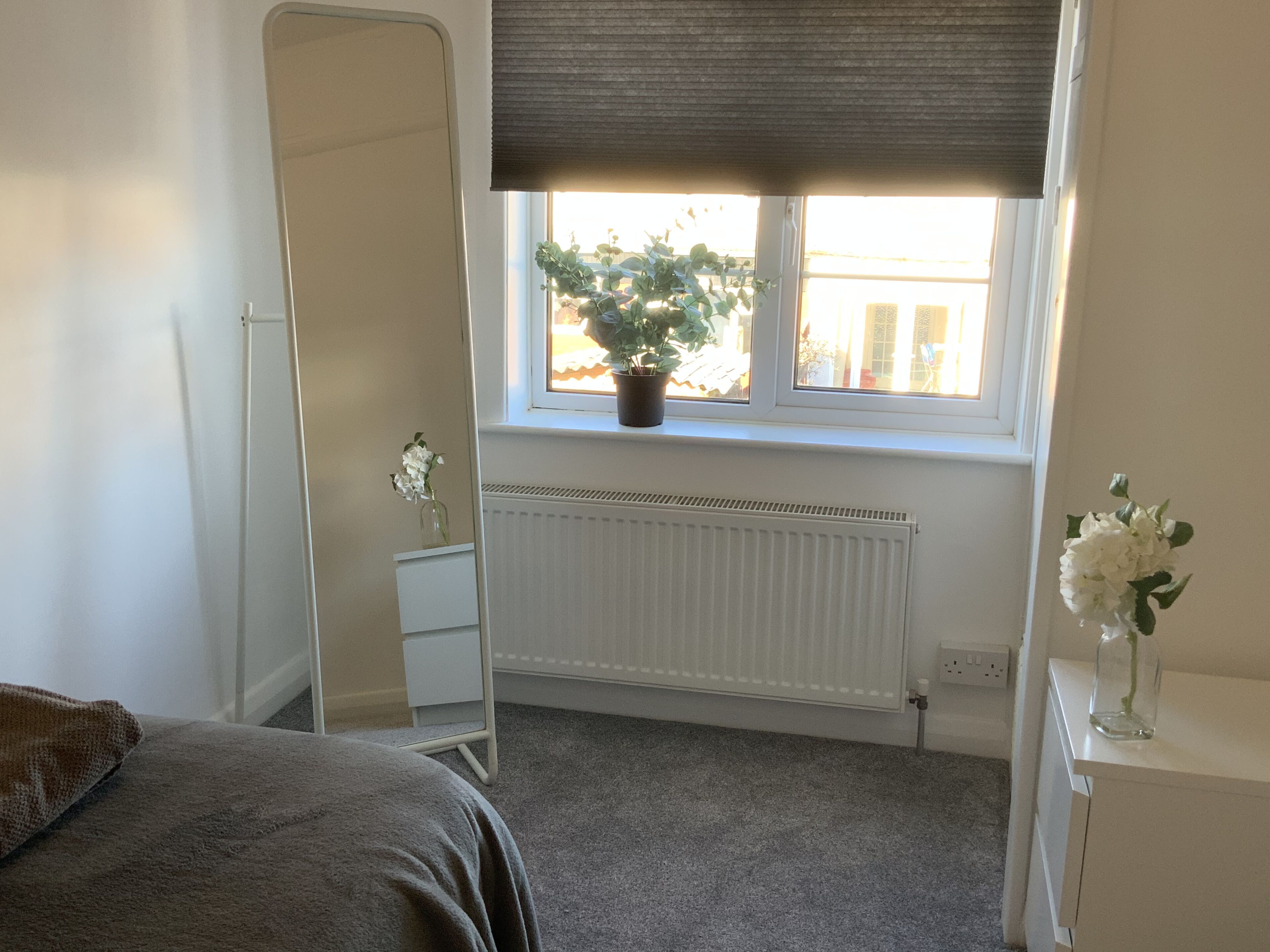 A bedroom with a large mirror, a potted plant on the windowsill, a radiator, and a bedside table with a vase of flowers.