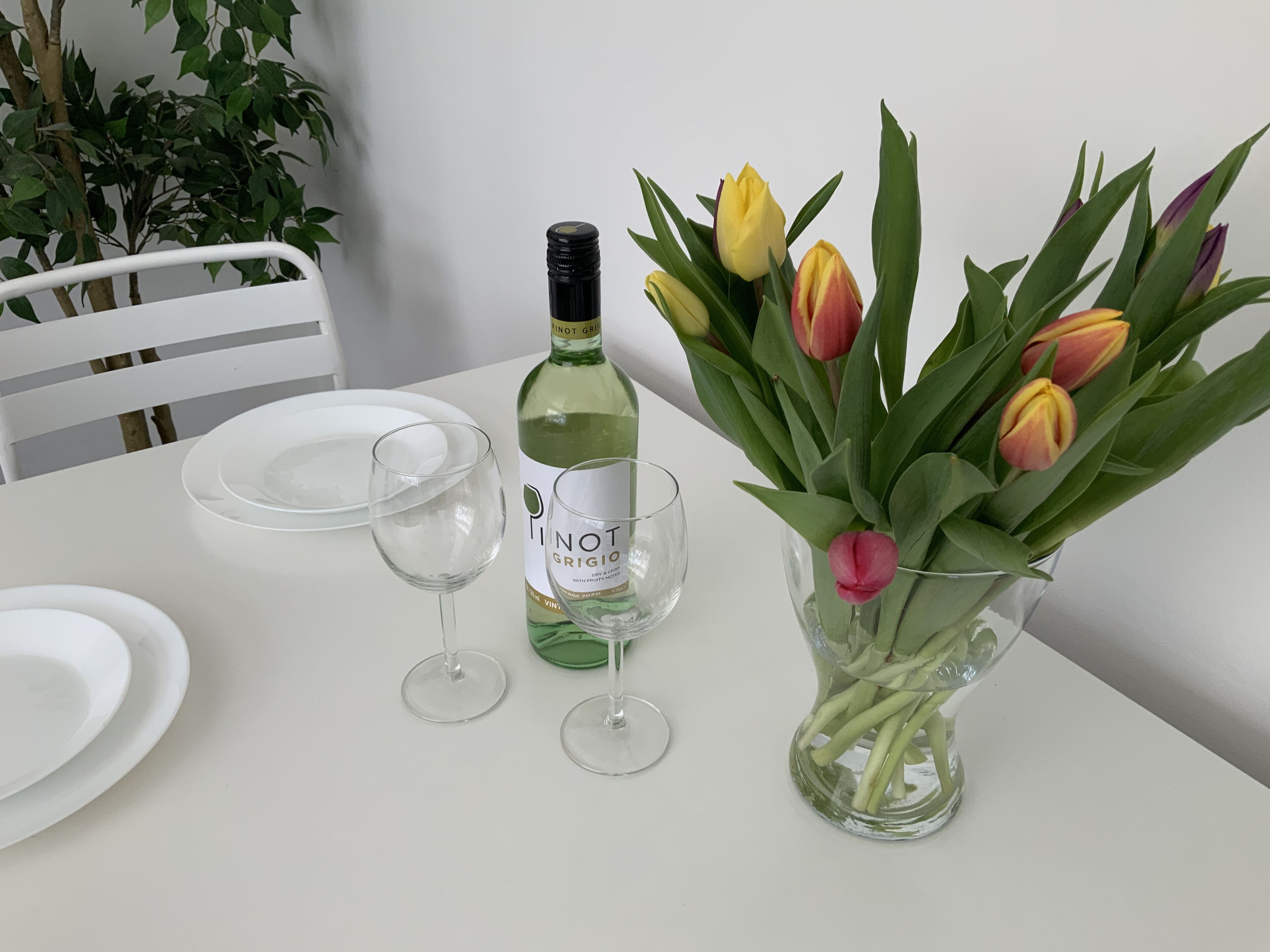 A dining table with a bottle of Pinot Grigio, two wine glasses, a vase with tulips, and white plates.