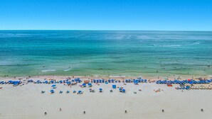 Una playa con numerosos sombrillas azules y sillas reclinables, una orilla arenosa y el océano al fondo.