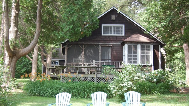 Une maison en bois rustique, dotée d’un porche, entourée d’une végétation luxuriante et agrémentée de trois chaises Adirondack.