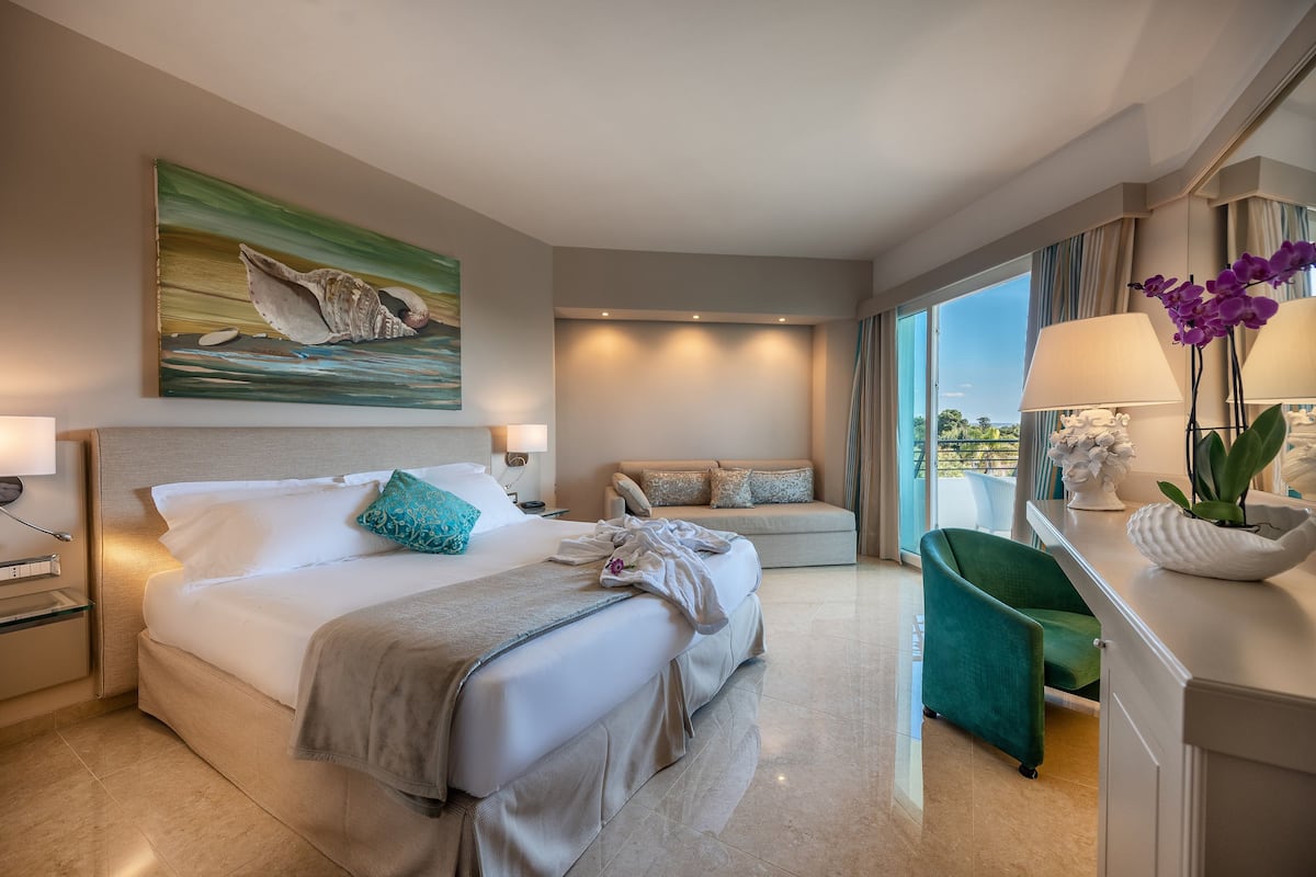 A modern hotel room with a large bed, a sofa, a desk with a lamp, and a view of the outdoors.
