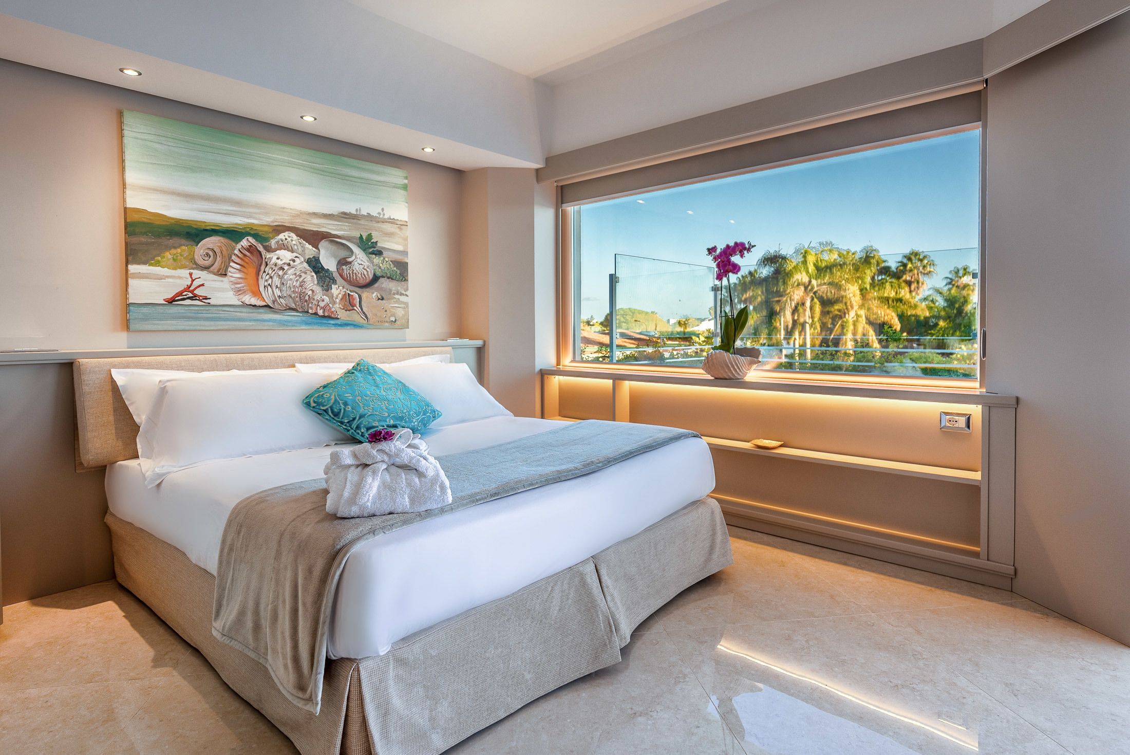 A modern bedroom with a large bed, a painting of seashells, a view of palm trees, and a window with a view of the outdoors.