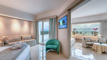 A modern hotel room with a sofa, a green armchair, a dining area with a table, and a TV mounted on the wall.