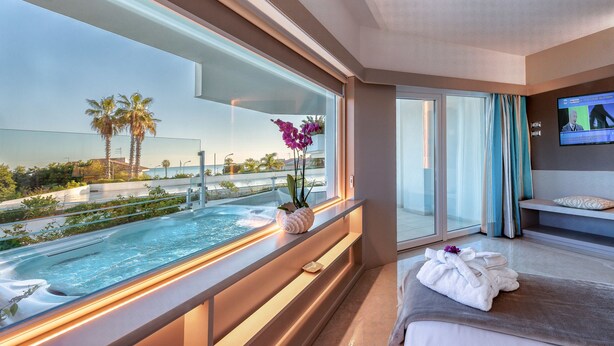 Habitación de hotel con un ventanal grande que da a una piscina y palmeras, un televisor en la pared y una camilla de masajes con toallas.