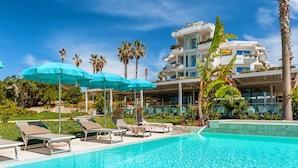 Zona de piscina con sillas de descanso y sombrillas, rodeada de palmeras y un edificio moderno con balcones.