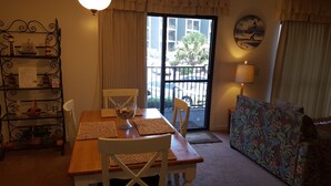 Uma área de refeições com uma mesa de madeira, cadeiras, uma tigela de petiscos, uma estante decorativa com objetos, uma janela com vista para edifícios e árvores, e um sofá com padrão floral.