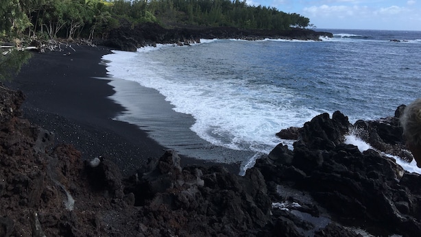 Un paisaje costero con playa de arena negra, costa rocosa y bosque denso.