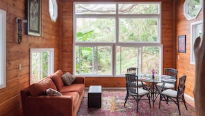Interior de una cabaña de madera con sofá, sillas, una mesa y grandes ventanas que dan a vistas de vegetación.
