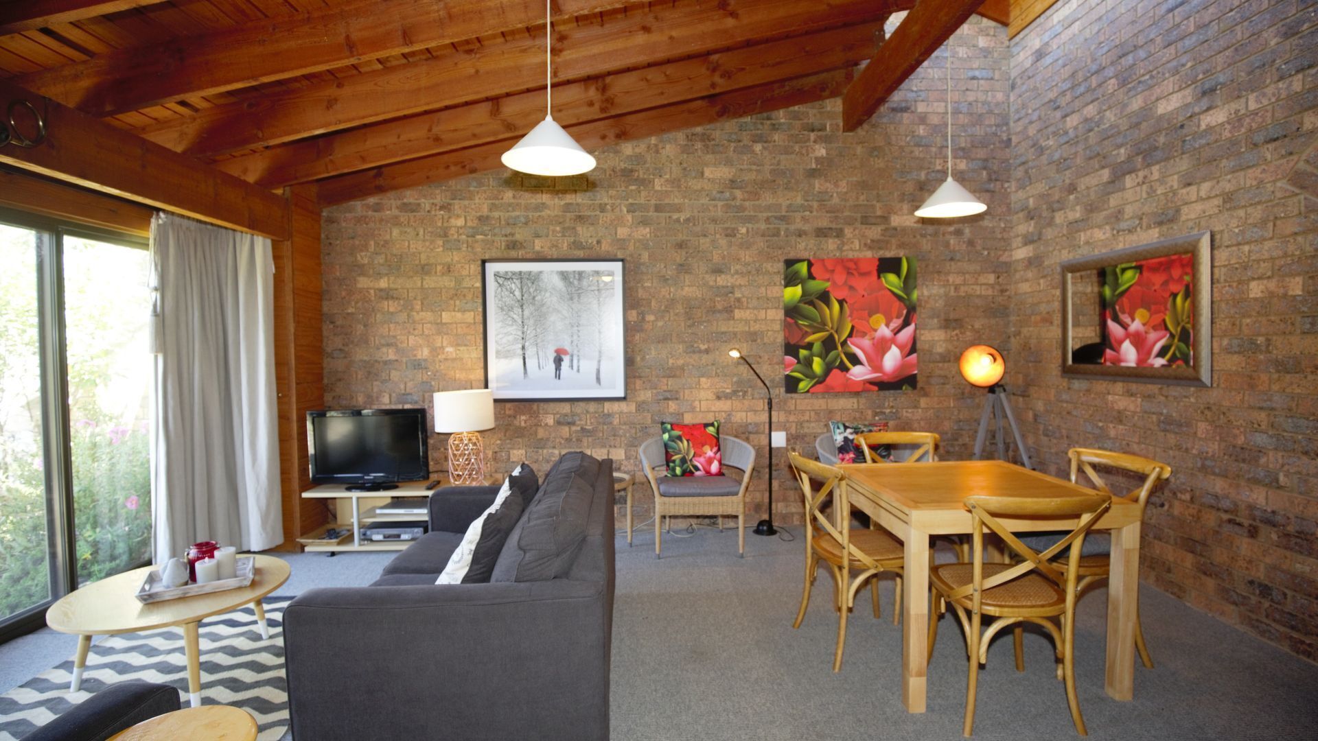 A cozy living room with a sofa, dining table, and chairs, a TV, and artwork on the brick walls.