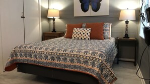 Een slaapkamer met een bed met decoratieve spreien, twee bedlampen, een schilderij van een vlinder aan de muur en een deur.