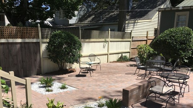 Een terras met bakstenen vloer, tuinmeubilair en een kleine tuin met planten en een decoratieve steen.