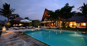 A swimming pool with lounge chairs and umbrellas, surrounded by palm trees and a building with a thatched roof.