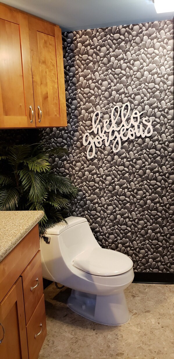 A bathroom with a toilet, wooden cabinets, a pebble wall, and a 'hello gorgeous' sign.
