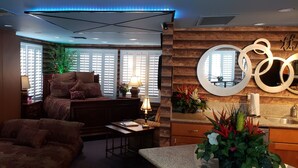A modern living room with a large bed, a sofa, a coffee table, and a dining area with a sink and decorative plants.