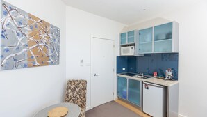 Cocina pequeña con microondas, lavamanos y armarios. Se observa una puerta con manija, una mesa pequeña con un tapete tejido y un cuadro abstracto colgado en la pared.