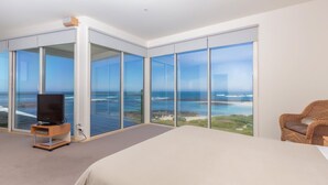 Una habitación de hotel moderna con una cama grande, un televisor y una silla de mimbre. La habitación tiene puertas de vidrio del suelo al techo que ofrecen vistas al océano.