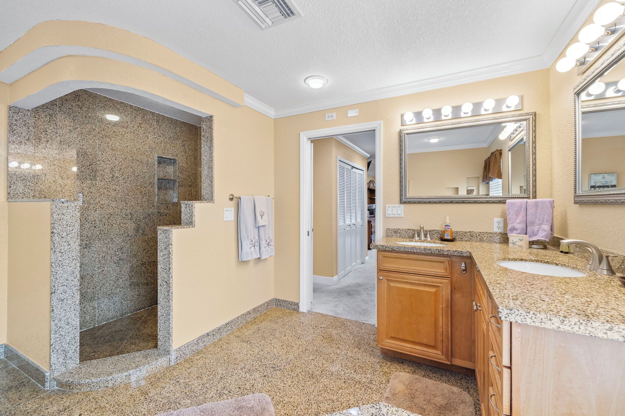 A bathroom with a granite shower, a vanity with a sink, and a large mirror with lights.